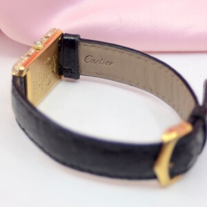 May include: A gold Cartier watch with a black leather band. The watch face is rectangular and has a diamond bezel. The band has the word 'Cartier' embossed on the back.