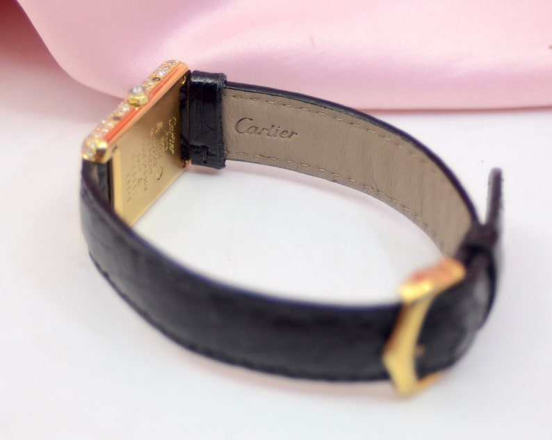 May include: A gold Cartier watch with a black leather band. The watch face is rectangular and has a diamond bezel. The band has the word 'Cartier' embossed on the back.