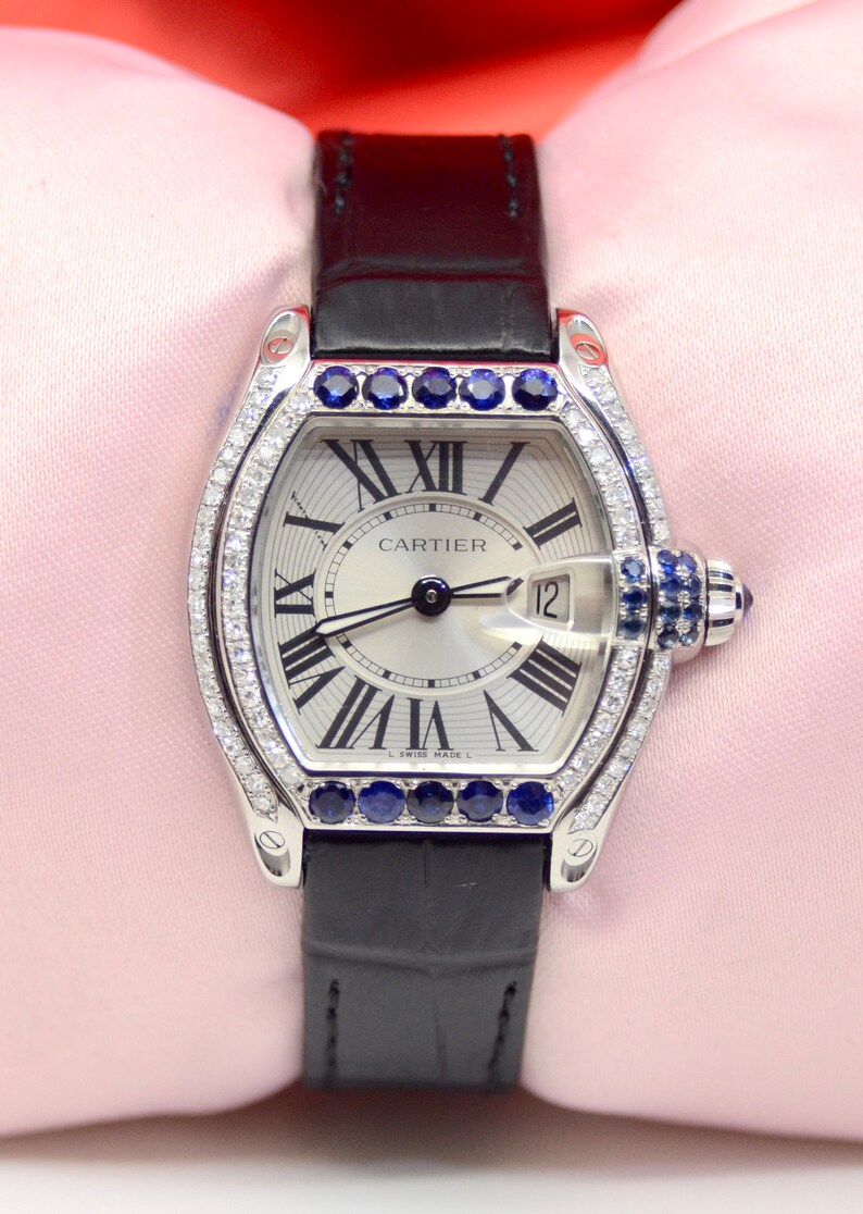 May include: A silver Cartier watch with a white face and black leather band. The watch has a diamond bezel and blue sapphire accents. The face of the watch has Roman numeral hour markers and a date window at the 12 o'clock position. The watch face also includes the text 'CARTIER' and 'Swiss Made'.