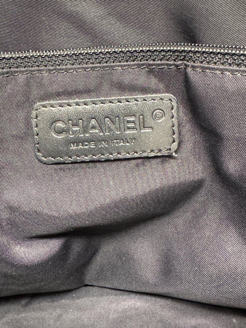 K&ouml;nnte beinhalten: Nahaufnahme eines schwarzen Lederetiketts mit dem gepr&auml;gten Wort "CHANEL" und dem Text "MADE IN ITALY". Das Etikett ist auf einem schwarzen Stoff befestigt, m&ouml;glicherweise die Innenseite einer Tasche. Oben ist ein schwarzer Rei&szlig;verschluss zu sehen.