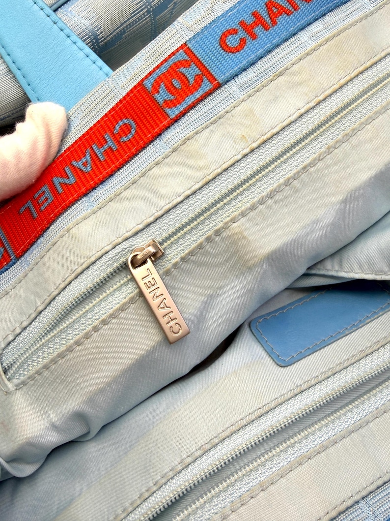 Puede incluir: Bolso de mano azul claro con una correa roja y azul con el nombre de la marca. El bolso tiene una cremallera plateada y un asa de cuero azul. El interior es azul claro.