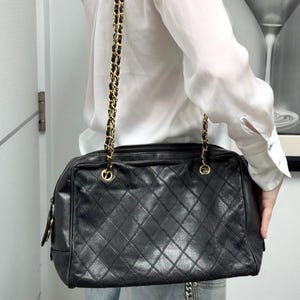 May include: Black quilted leather handbag with a gold chain strap. The bag has a rectangular shape and a diamond pattern. The chain strap is a combination of gold metal and black leather. The bag is a classic style.