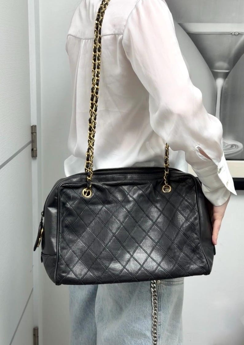 May include: Black quilted leather handbag with a gold chain strap. The bag has a rectangular shape and a diamond pattern. The chain strap is a combination of gold metal and black leather. The bag is a classic style.
