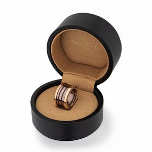 May include: A rose gold ring with black and pink bands, presented in an open black jewellery box with a tan interior. The brand name "BVLGARI" is visible inside the box.