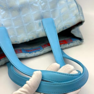 Puede incluir: Bolso tote azul claro con un patr&oacute;n acolchado y asas de cuero azul claro. El bolso presenta un patr&oacute;n de logotipo repetido y un forro interior rojo y azul. Las asas est&aacute;n sujetas por una mano enguantada.