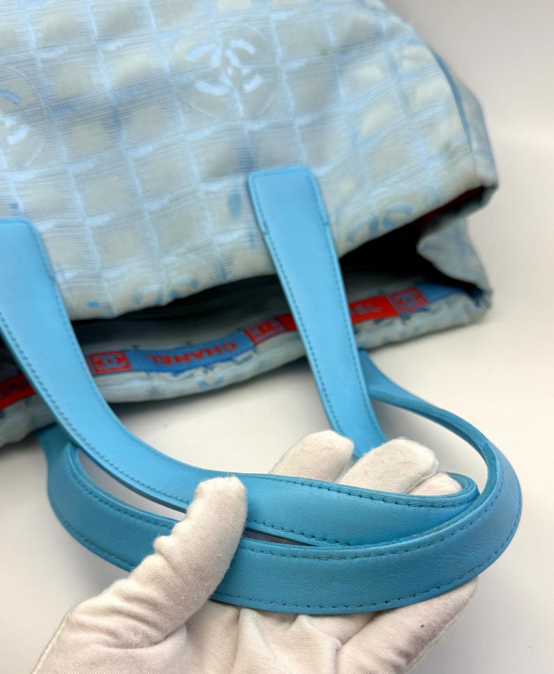 Puede incluir: Bolso tote azul claro con un patr&oacute;n acolchado y asas de cuero azul claro. El bolso presenta un patr&oacute;n de logotipo repetido y un forro interior rojo y azul. Las asas est&aacute;n sujetas por una mano enguantada.