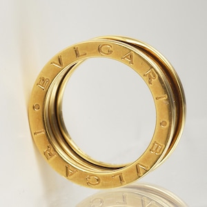 May include: A gold-colored ring with three interlocking bands. The outer bands feature the engraved word "BVLGARI". The ring is displayed on a white background, highlighting its polished surface and design.