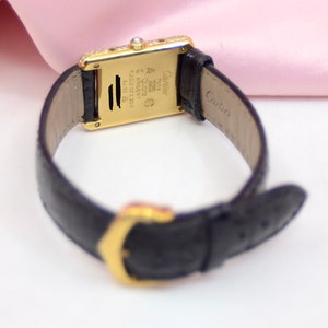 May include: A gold Cartier watch with a black leather band. The watch face is rectangular and has a diamond bezel. The back of the watch has the Cartier logo and the words 'Swiss Made' and '18K 750'.
