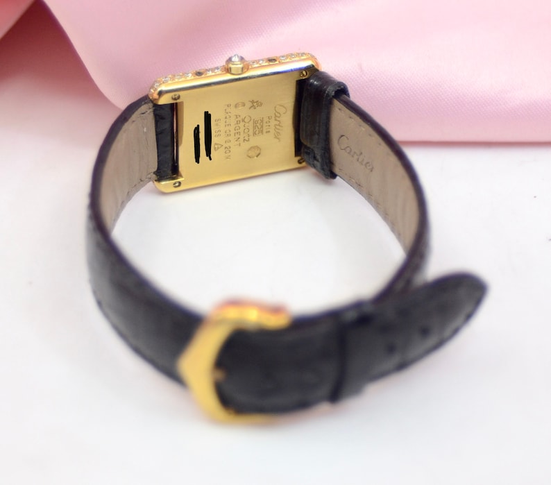 May include: A gold Cartier watch with a black leather band. The watch face is rectangular and has a diamond bezel. The back of the watch has the Cartier logo and the words 'Swiss Made' and '18K 750'.