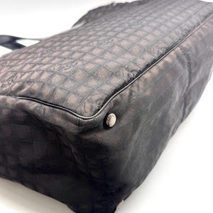 K&ouml;nnte beinhalten: Dunkelbraune Tragetasche mit sich wiederholendem quadratischem Muster. Die Tasche hat eine schwarze Umrandung und einen silbernen Knopf. Das Material scheint ein gewebter Stoff zu sein. Die Tasche befindet sich auf wei&szlig;em Hintergrund.