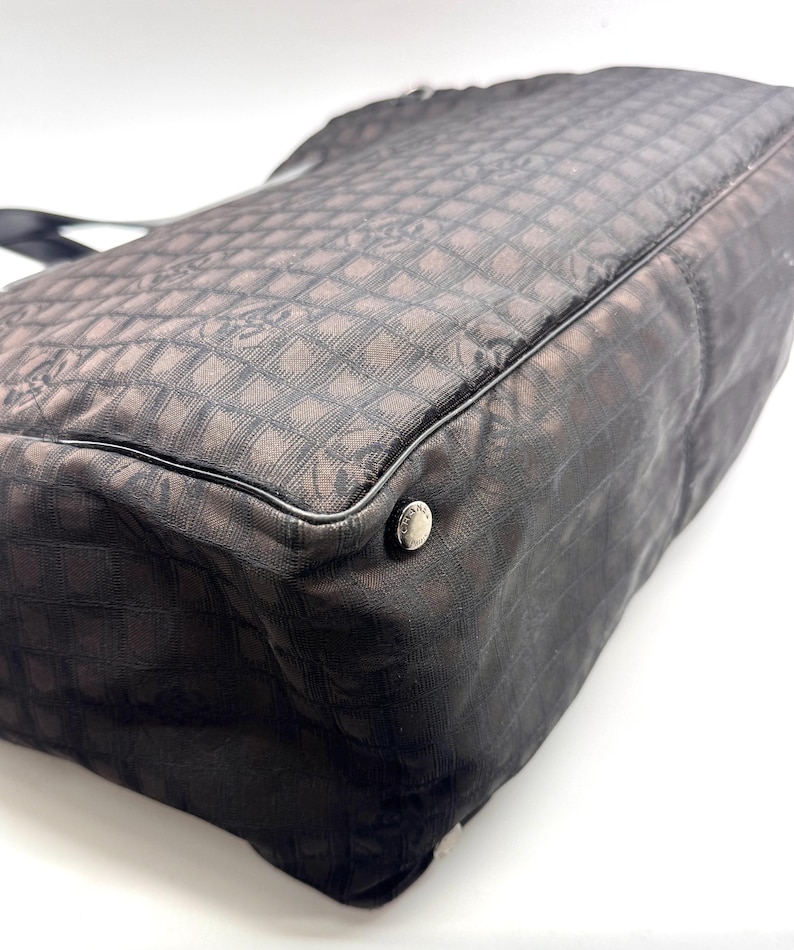 K&ouml;nnte beinhalten: Dunkelbraune Tragetasche mit sich wiederholendem quadratischem Muster. Die Tasche hat eine schwarze Umrandung und einen silbernen Knopf. Das Material scheint ein gewebter Stoff zu sein. Die Tasche befindet sich auf wei&szlig;em Hintergrund.