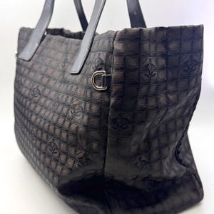 K&ouml;nnte beinhalten: Eine braune Tragetasche mit einem sich wiederholenden quadratischen Muster und einem Logo-Design. Die Tasche hat schwarze Ledergriffe und silberfarbene Hardware. Die Tasche besteht aus einem gewebten Stoff.