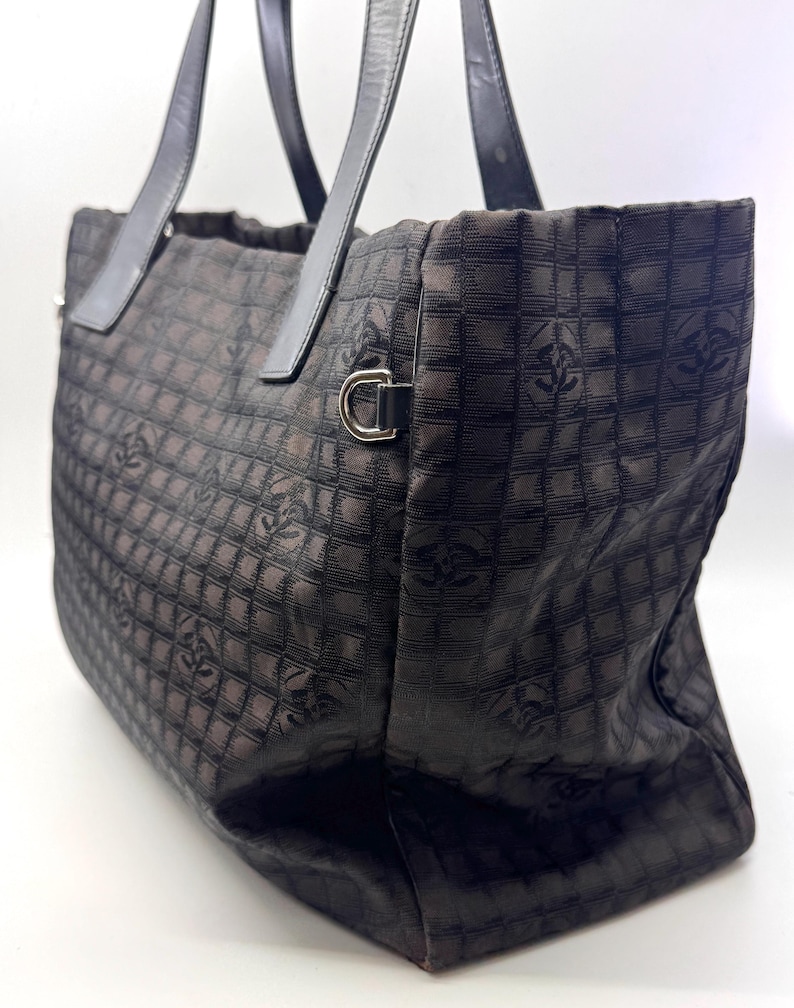 K&ouml;nnte beinhalten: Eine braune Tragetasche mit einem sich wiederholenden quadratischen Muster und einem Logo-Design. Die Tasche hat schwarze Ledergriffe und silberfarbene Hardware. Die Tasche besteht aus einem gewebten Stoff.