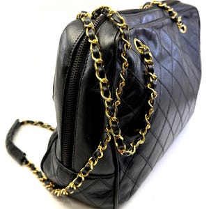 May include: Black quilted leather handbag with gold-tone chain straps. The bag features a diamond pattern and a top zipper closure. The chain strap is interwoven with black leather. A classic and elegant accessory.