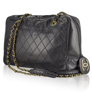 May include: Black quilted leather handbag with gold-tone chain straps. The bag features a diamond pattern and a zipper closure. A gold-tone logo charm hangs from the side. The bag has a rectangular shape.