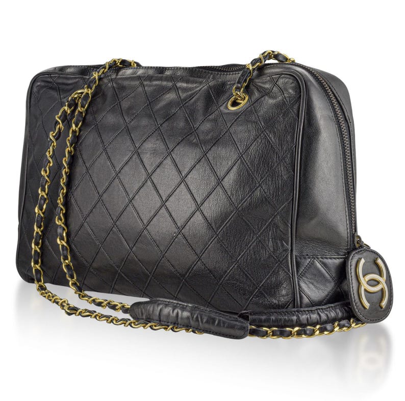May include: Black quilted leather handbag with gold-tone chain straps. The bag features a diamond pattern and a zipper closure. A gold-tone logo charm hangs from the side. The bag has a rectangular shape.