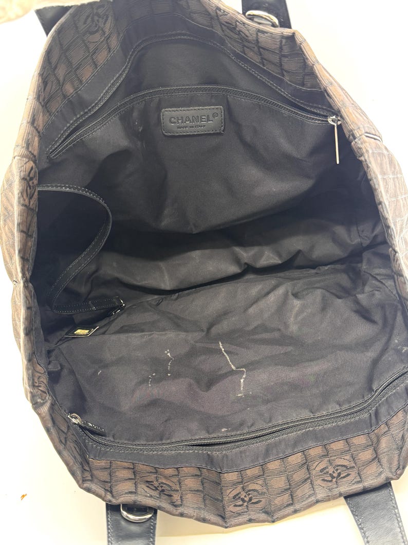 K&ouml;nnte beinhalten: Offene Ansicht einer braunen und schwarzen Tragetasche mit sich wiederholendem Muster. Die Tasche hat schwarze Griffe und ein schwarzes Innenfutter mit mehreren F&auml;chern. Ein kleines Lederetikett mit dem Markennamen ist innen sichtbar.