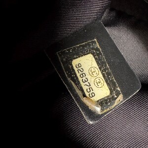 K&ouml;nnte beinhalten: Nahaufnahme eines schwarzen Stoffes mit einem rechteckigen Etikett. Das Etikett ist cremefarben mit der Nummer "9263759" und zwei ineinandergreifenden "C"-Logos. Das Etikett ist in klarem Kunststoff eingeschlossen.