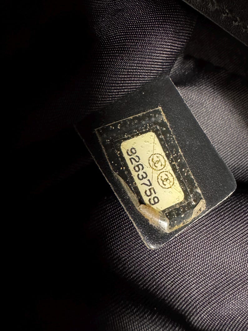 K&ouml;nnte beinhalten: Nahaufnahme eines schwarzen Stoffes mit einem rechteckigen Etikett. Das Etikett ist cremefarben mit der Nummer "9263759" und zwei ineinandergreifenden "C"-Logos. Das Etikett ist in klarem Kunststoff eingeschlossen.