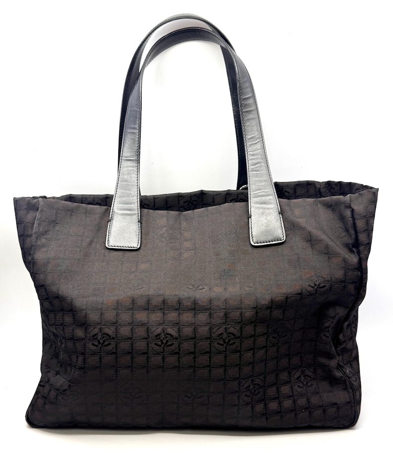 K&ouml;nnte beinhalten: Eine dunkelbraune Tragetasche mit sich wiederholendem Muster und silberfarbenen Griffen. Die Tasche hat eine rechteckige Form und ein strukturiertes Design. Die Griffe sind mit silberfarbenen Akzenten an der Tasche befestigt. Die Tasche besteht aus einem gewebten Material.