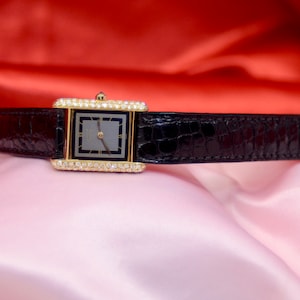 May include: A gold watch with a black leather band and diamond accents around the face. The watch face is rectangular and has a black background with gold hands and hour markers.