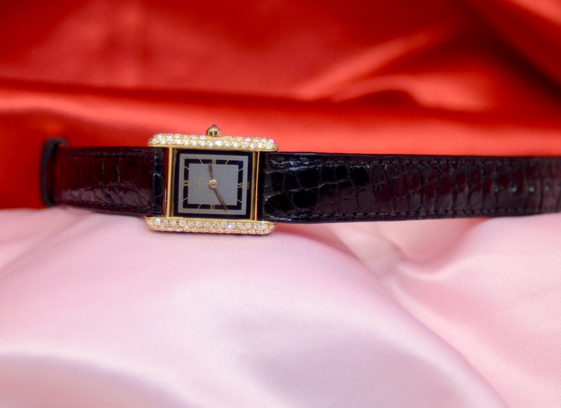 May include: A gold watch with a black leather band and diamond accents around the face. The watch face is rectangular and has a black background with gold hands and hour markers.
