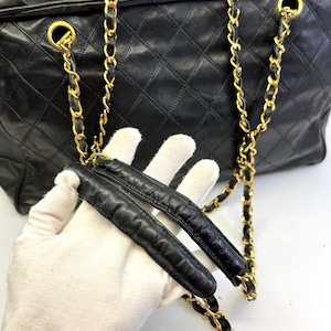 May include: Black quilted leather handbag with gold-tone chain straps. The bag features a diamond pattern and a zipper closure. The chain straps are intertwined with black leather. The bag is held by a gloved hand.