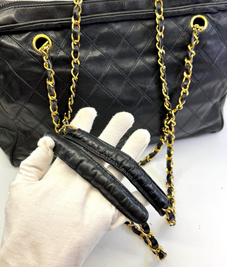 May include: Black quilted leather handbag with gold-tone chain straps. The bag features a diamond pattern and a zipper closure. The chain straps are intertwined with black leather. The bag is held by a gloved hand.