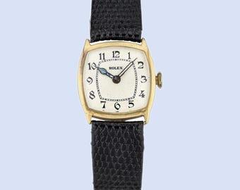 May include: A vintage Rolex wristwatch with a gold-toned rectangular case and a cream-colored face. The watch features black Arabic numerals and hands, with the brand name "ROLEX" displayed. It has a black leather strap.