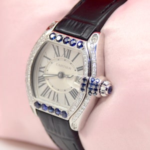 May include: A silver Cartier watch with a white face and black leather band. The watch is set with diamonds and sapphires around the bezel. The watch face has Roman numeral hour markers and the word 'CARTIER' printed at the top.