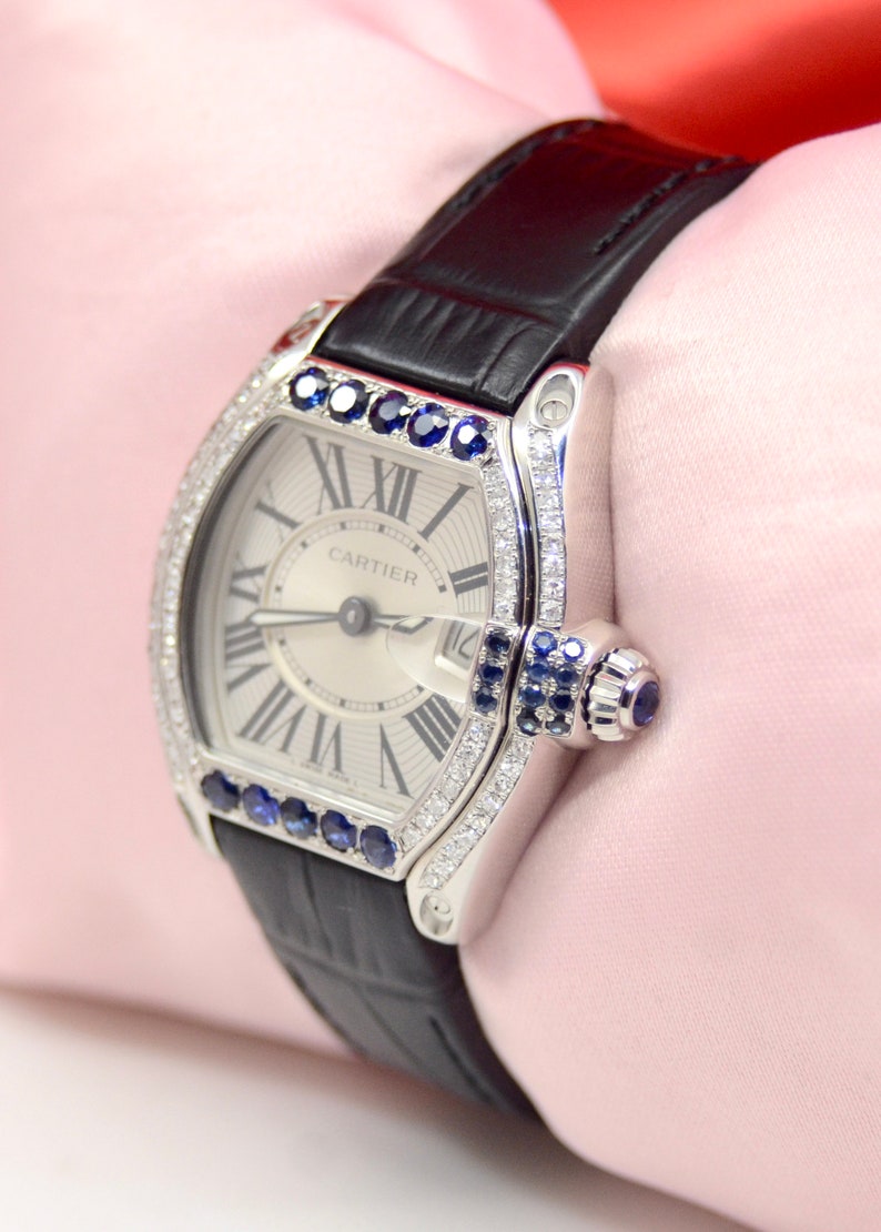 May include: A silver Cartier watch with a white face and black leather band. The watch is set with diamonds and sapphires around the bezel. The watch face has Roman numeral hour markers and the word 'CARTIER' printed at the top.