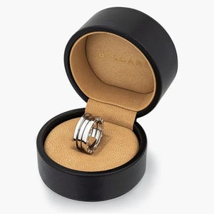 May include: A silver ring with three bands sits in an open, black, round jewellery box with a tan interior. The ring's polished metal finish is the focal point. The box is open, displaying the ring's design.
