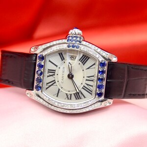 May include: A silver Cartier watch with a white face and black leather band. The watch has a diamond bezel and blue sapphire accents. The face of the watch has Roman numeral hour markers and the words 'Cartier' printed on it.