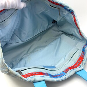 Puede incluir: Bolso tote azul claro con estampado de cuadros y asas de cuero azul. El interior es azul claro con m&uacute;ltiples compartimentos. Ribete rojo y azul con la palabra "CHANEL" visible en el borde superior.