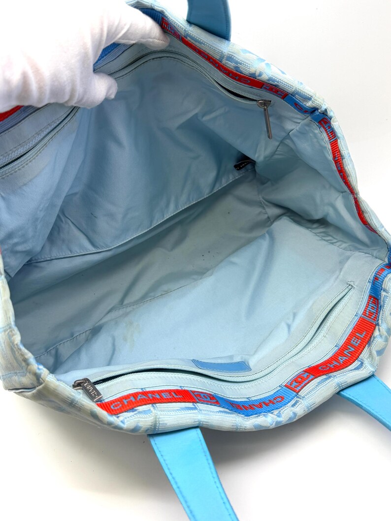 Puede incluir: Bolso tote azul claro con estampado de cuadros y asas de cuero azul. El interior es azul claro con m&uacute;ltiples compartimentos. Ribete rojo y azul con la palabra "CHANEL" visible en el borde superior.