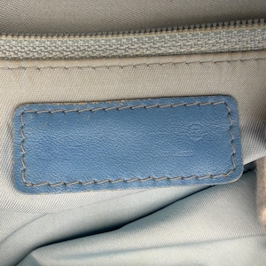 Puede incluir: Primer plano de una etiqueta de cuero azul claro con un borde cosido. La etiqueta es rectangular y est&aacute; colocada sobre un fondo de tela azul claro. Una cremallera plateada es visible en la parte superior.