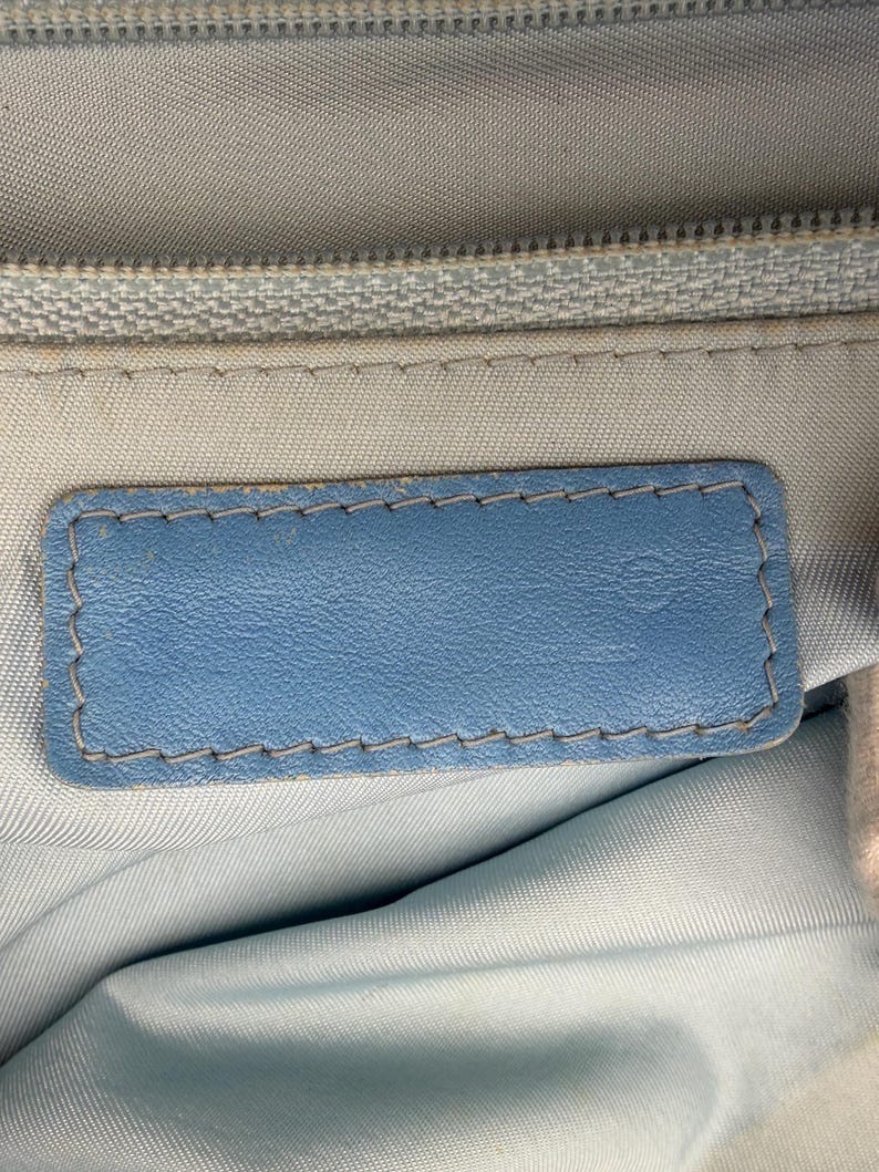 Puede incluir: Primer plano de una etiqueta de cuero azul claro con un borde cosido. La etiqueta es rectangular y est&aacute; colocada sobre un fondo de tela azul claro. Una cremallera plateada es visible en la parte superior.