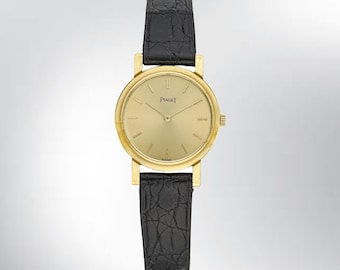 May include: A gold-toned Piaget watch with a black leather band. The watch face is round with gold hands and markers. The band is textured. The watch is a classic timepiece.
