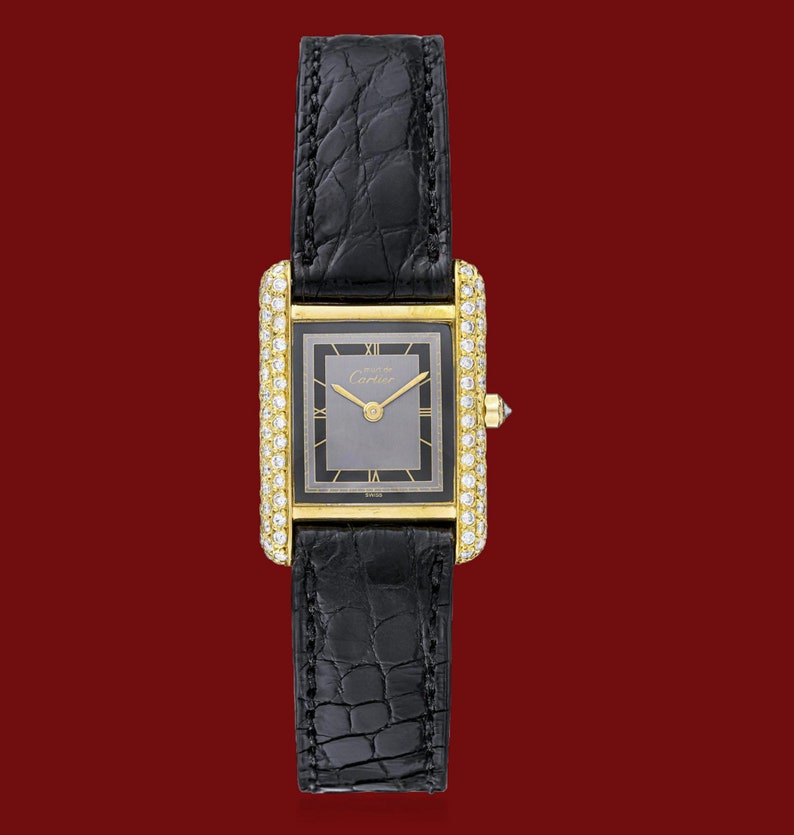 May include: A gold wristwatch with a black leather band and a diamond bezel. The watch face is gray with black Roman numeral hour markers and black hands. The watch face also has the text 'Cartier' printed on it.