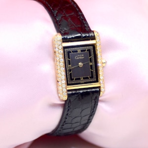 May include: A gold Cartier watch with a black face and diamond bezel. The watch has a black alligator strap and is set against a pink background.