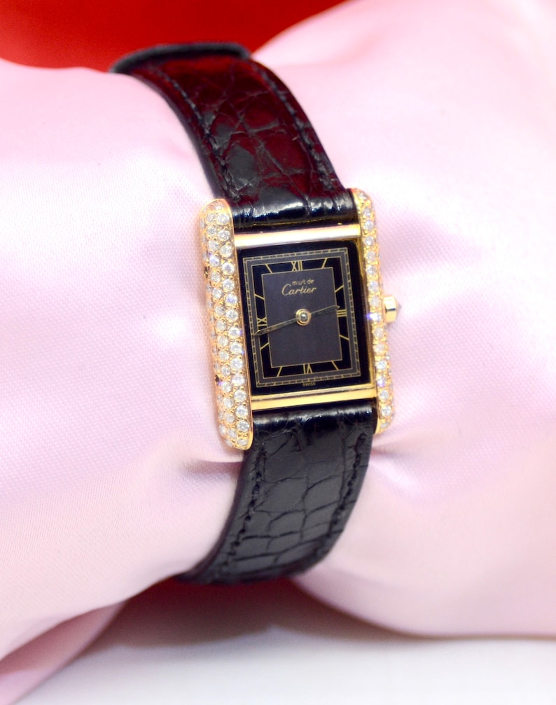 May include: A gold Cartier watch with a black face and diamond bezel. The watch has a black alligator strap and is set against a pink background.