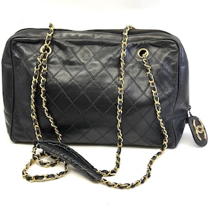 May include: Black quilted leather handbag with gold-tone chain straps. The rectangular bag features a diamond pattern and a zipper closure. A small, round charm with a logo hangs from the side.