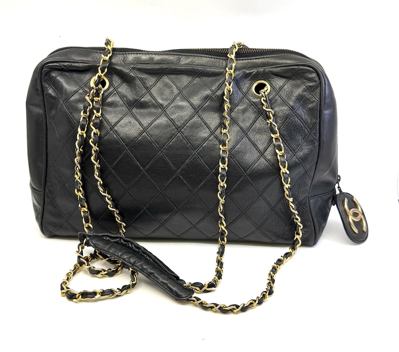 May include: Black quilted leather handbag with gold-tone chain straps. The rectangular bag features a diamond pattern and a zipper closure. A small, round charm with a logo hangs from the side.
