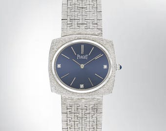 May include: A silver-toned Piaget wristwatch featuring a round, deep blue dial with silver hands and three diamond hour markers. The watch has a textured, brushed metal case and a matching integrated link bracelet with a distinct geometric pattern.