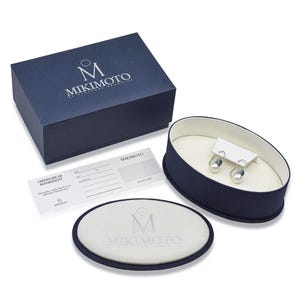 May include: An open navy blue jewelry box with a pair of pearl earrings displayed inside. The earrings are on a white card. A matching navy blue box and a certificate of authenticity are also present. The Mikimoto logo is visible on the box and the card.