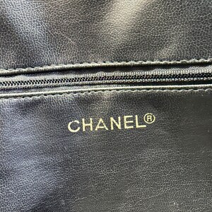 May include: Close-up of a black leather item with a textured surface. A zipper is visible near the top. The word "CHANEL" is printed in gold, with the registered trademark symbol.