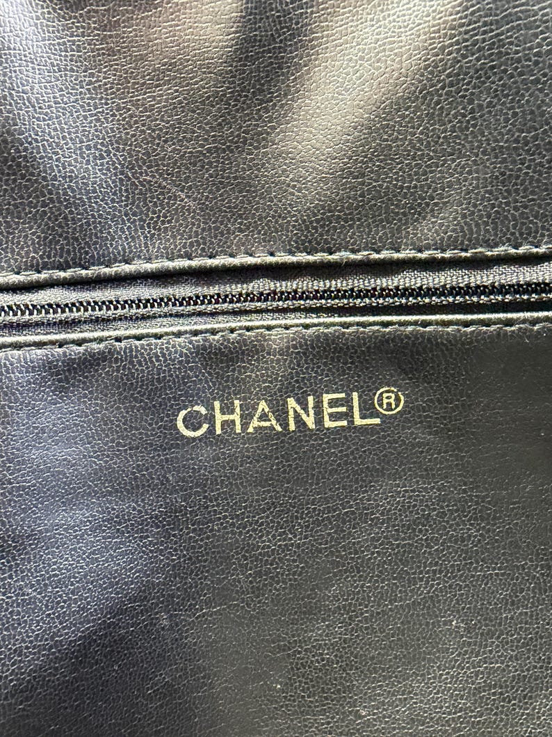 May include: Close-up of a black leather item with a textured surface. A zipper is visible near the top. The word "CHANEL" is printed in gold, with the registered trademark symbol.