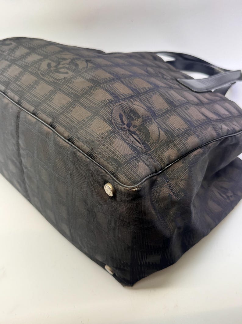 May include: A black tote bag with a repeating pattern of interlocking Cs. The bag has a rectangular shape with a flat bottom and a top handle. The bag's corners have silver-colored metal studs.