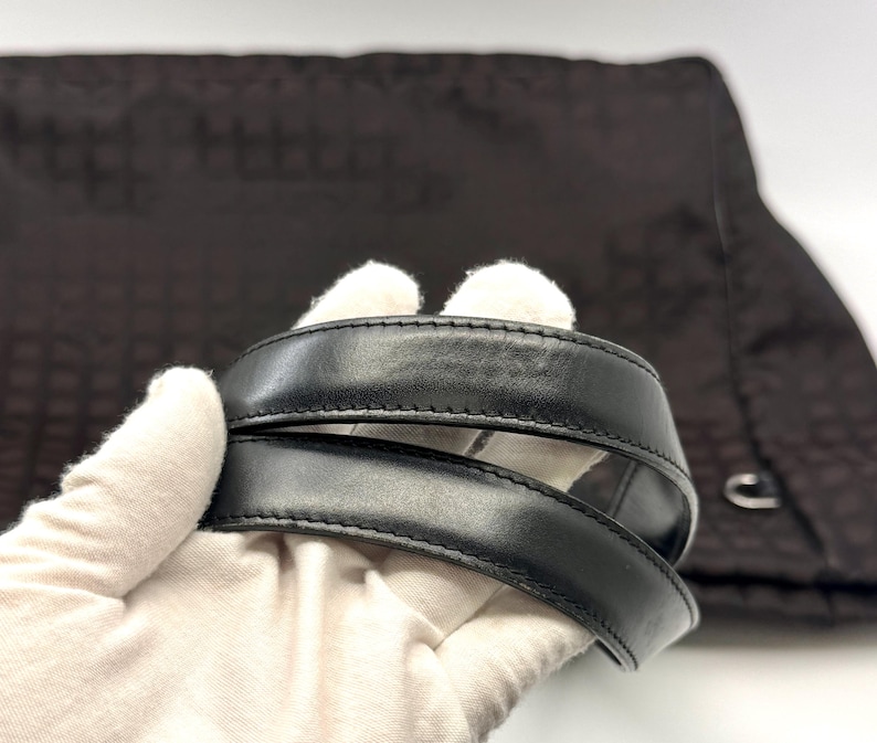 K&ouml;nnte beinhalten: Nahaufnahme eines schwarzen Lederhandtaschengriffs. Der Griff ist dick und gen&auml;ht, mit einer glatten, gl&auml;nzenden Oberfl&auml;che. Der Hintergrund ist ein dunkelbrauner Stoff mit einem dezenten Muster. Die Tasche scheint eine Tragetasche oder eine Umh&auml;ngetasche zu sein.