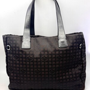 K&ouml;nnte beinhalten: Dunkelbraune Tragetasche mit sich wiederholendem Muster. Die Tasche hat zwei schwarze Ledergriffe und silberfarbene Metallringe an den Seiten. Das Material der Tasche hat einen dezenten Glanz.